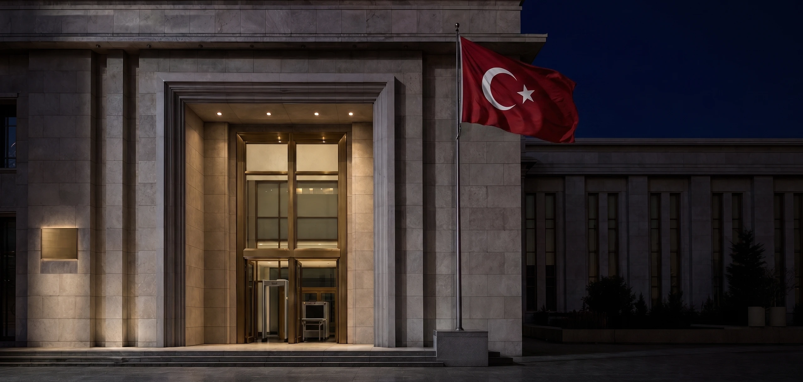Akşam saatinde bir kamu binasının aydınlatılmış giriş cephesi ve yanında Türk bayrağı.